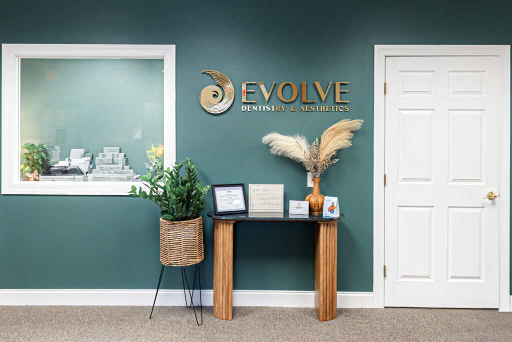 The main lobby of Evolve Dentistry & Aesthtics, a small table displaying certiications and some potted plants decoarting the room. To the right of the tbale, there is a door, where a cosmetic dentist in Craven County, will take pateints to our treatment room. 